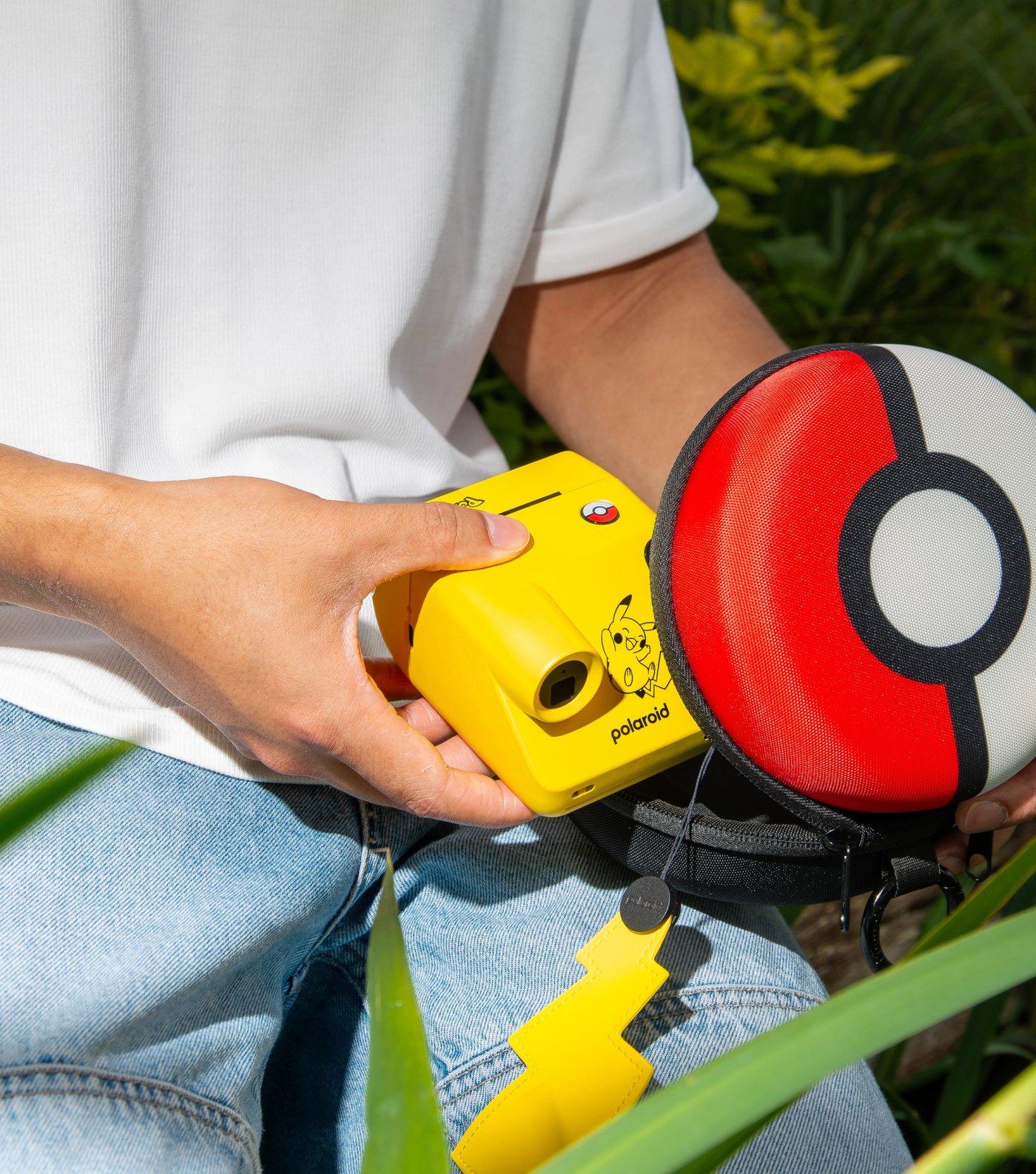 Un Polaroid Go Génération 3 édition Pikachu avec sa dragonne en forme d'éclair, à côté de l'étui en forme de Poké Ball